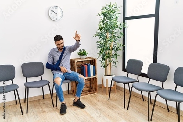 Fototapeta Young arab man suffering sitting on chair with arm sling and finger raised up at clinic waiting room