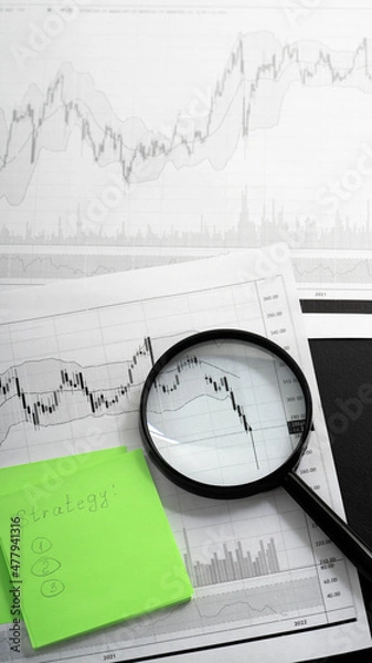 Fototapeta Financial instrument movement charts on white sheet of paper, magnifying glass on rebound chart, pen and sheets for notes lie on table. Preparing an entry strategy in a stock.