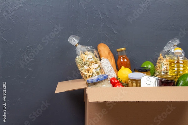 Obraz Closeup view of donation box with different food donations on gray wall background with copy space - pasta, fresh vegatables, canned food, baguette, cooking oil. Food bank, food delivery concept