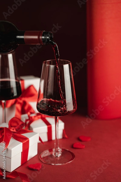 Fototapeta Two glasses and bottle of red wine, hearts, gifts boxes for Valentine day. Modern Still life. Red background. Greeting card for Valentine's Day