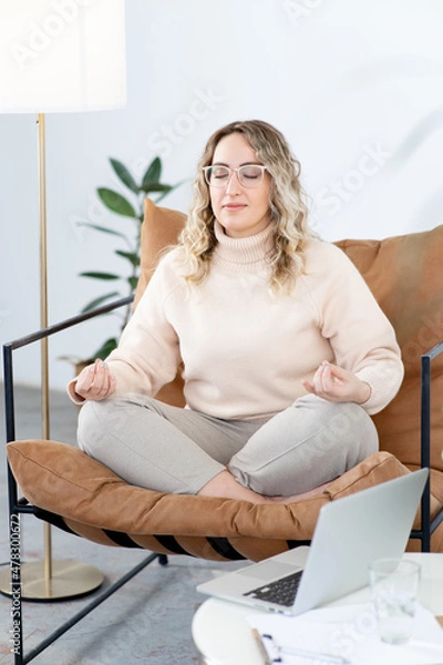 Fototapeta Blonde woman meditates sitting in a chair in the lotus position. Rest from the hard psychological work of a psychotherapist. There is an open laptop on table. There is a large floor lamp nearby.