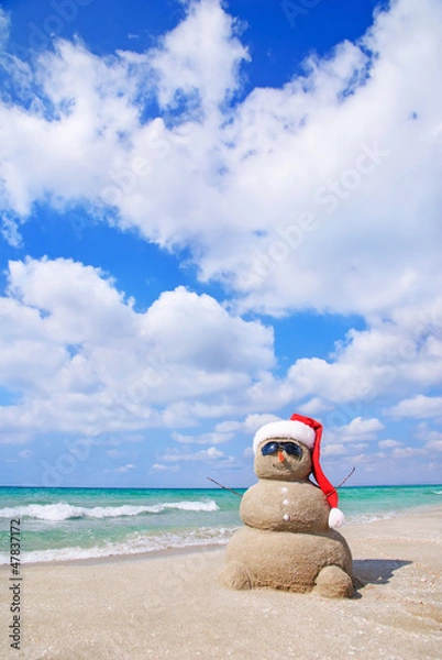 Fototapeta Smiling sandy snowman in red santa hat on the sea beach.