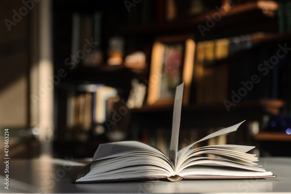 Obraz There is an open book on the table.  The background is bookshelves illuminated by the sun.