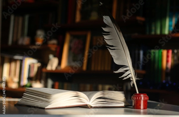 Obraz An open book. Next to it is an inkwell with a quill pen. Background - bookshelves illuminated by the sun.
