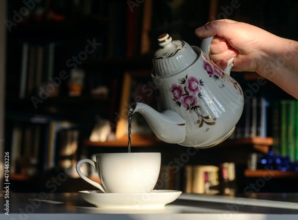 Obraz There is a teapot in a woman's hand. She pours tea into a cup.  Background - bookshelves illuminated by the sun.