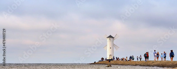 Fototapeta Tourists walk on the pier to a lighthouse on the Baltic Sea in Swinoujscie in Poland. With blurred cloudy sky in the background.