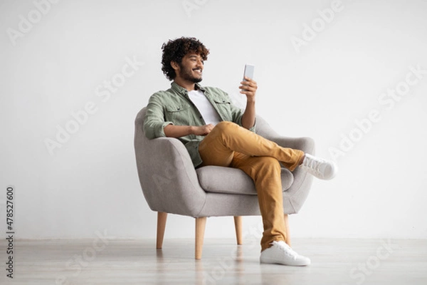 Fototapeta Cheerful young indian man chatting with girlfriend, using smartphone
