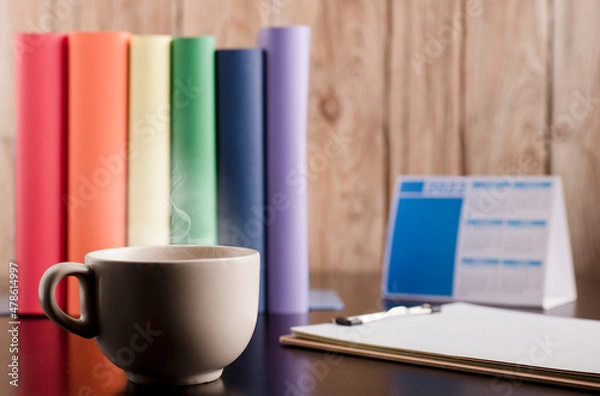 Fototapeta Cup of coffee or tea with hot steam in the morning, books with lgtbi flag colors, on the desk.