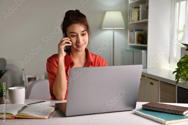 Fototapeta Beautiful young  Asian woman work at home with laptop and talking on smartphone and smiling. Female working on computer and using cell mobile phone while sitting at home