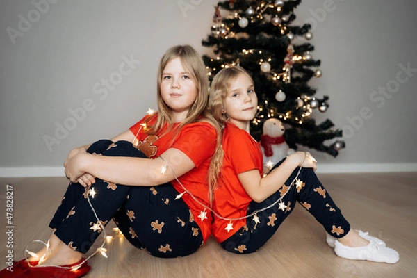 Fototapeta Teenage sisters in red holiday T-shirts sitting at the Christmas tree and celebrating the New Year