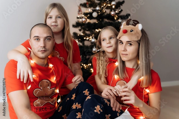 Fototapeta A family in red holiday T-shirts sitting at the Christmas tree and celebrating the New Year