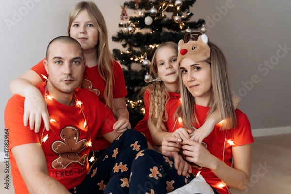 Fototapeta A family in red holiday T-shirts sitting at the Christmas tree and celebrating the New Year