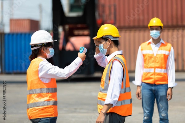 Fototapeta female manager measures temperature with a thermometer for employees before entering a container yard. import and export concept. Professional engineering team.