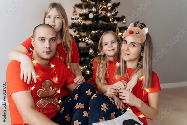 Fototapeta A family in red holiday T-shirts sitting at the Christmas tree and celebrating the New Year