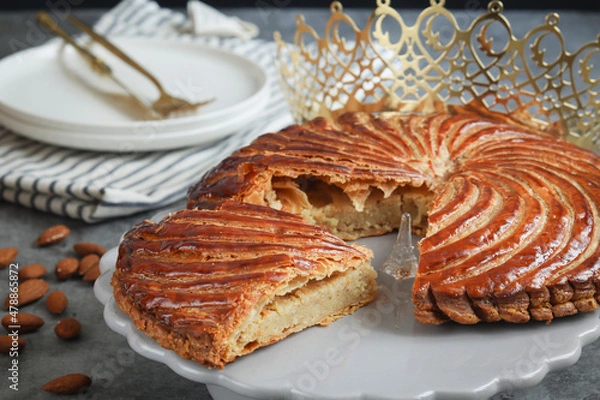 Fototapeta A traditional French galette des rois on a plate with crown, with a cut piece and its fève in a middle. Made puff pastry and creamy almond filling roll in circle shape. It's usually served on Epiphany