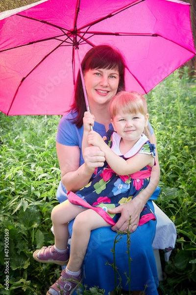 Fototapeta Happy young mother with funny little kid girl 3-4 year old walk outdoors with umbrella in a sunny summer or spring day during rain. Motherhood