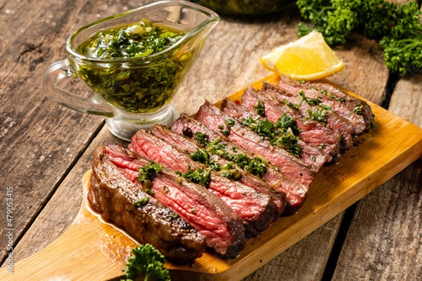 Fototapeta Juicy slices of chopped beef steak with aromatic chimichuri sauce on a wooden table.