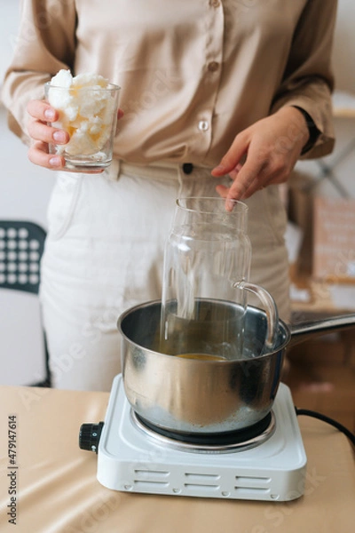 Fototapeta Vertical front view of female artisan adding white dry soy wax in glass jar into pot of boiling water for creating candle building mixture. Process of making handmade natural candle at workshop.