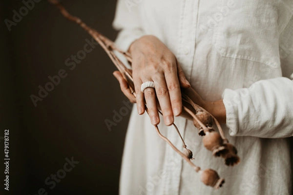 Obraz A woman is wearing a white cotton macrame ring while she is holding a dried poppy bunch. She is wearing white linen blouse. Copy space image. Sustainable fashion trendy accessory. 