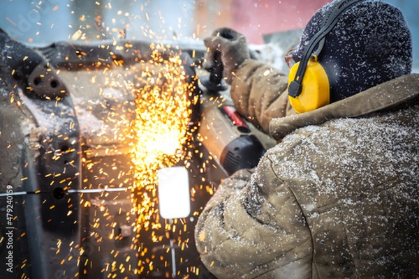 Fototapeta Worker in dirty clothes saws metal in winter in snowfall