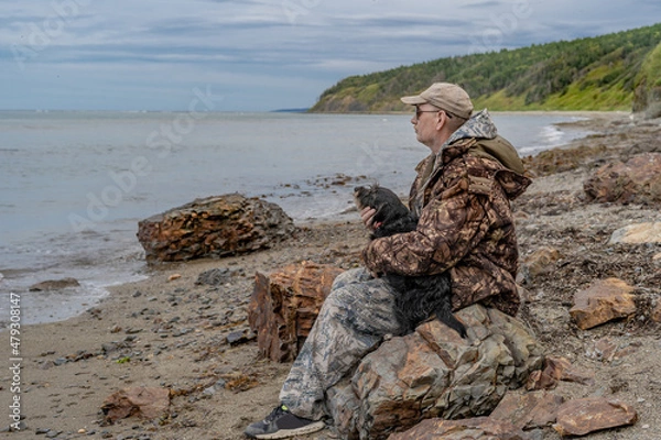 Fototapeta A pensive elderly man by the sea sits on a stone with a dog, thinking about the meaning of life.