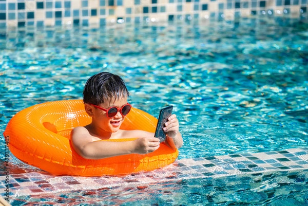 Obraz Asian boy swim and play game from mobile phone at the swimming pool in summer