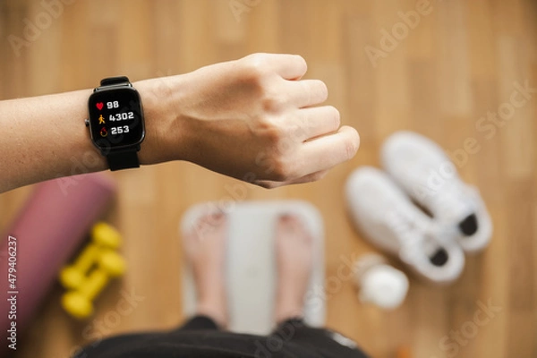 Fototapeta Looking at fitness tracker displaying heart rate, steps count and burnt calories while standing on scales, first person view. Smart watch on female hand, point of view, concept of staying fit at home