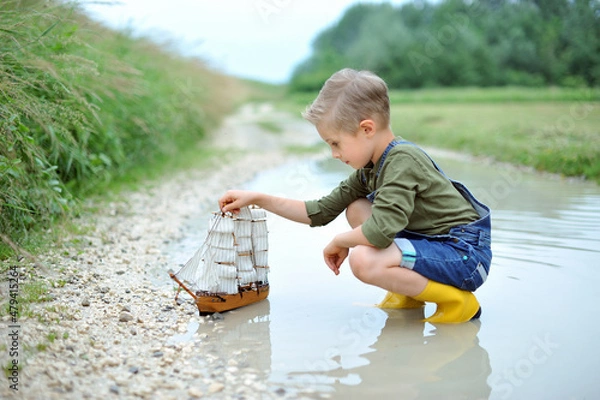 Fototapeta A boy, 7 years old, is playing with a small sailing ship in a puddle after the rain.