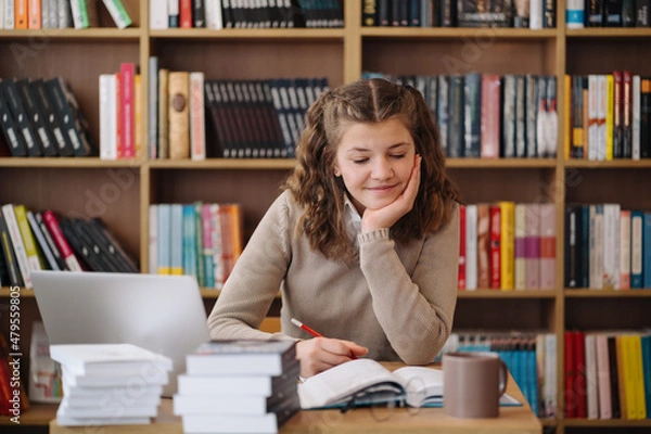 Obraz Attractive happy young girl student studying at the college library, sitting at the desk, using laptop computer, having video chat