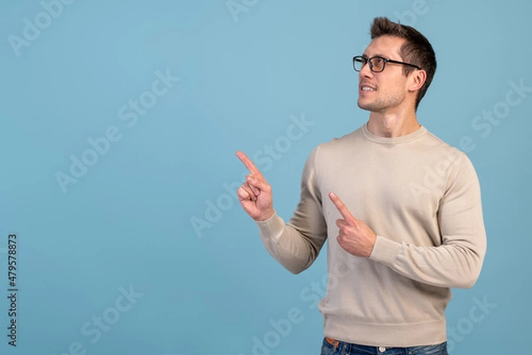 Fototapeta Advertise here. Positive bearded man pointing finger away paying your attention at empty space for advertisement. Indoor studio shot isolated on blue background