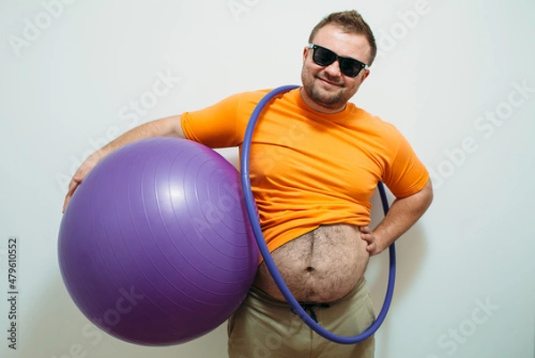 Fototapeta Funny image of a fat guy holding a hula hoop and exercise ball demonstrates his big belly. Fitness time. Time to lose weight concept.