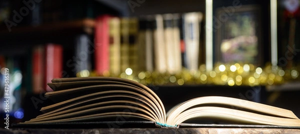 Obraz The open book, illuminated by the sun, lies on the table. Bookshelf background and blurry color spots.