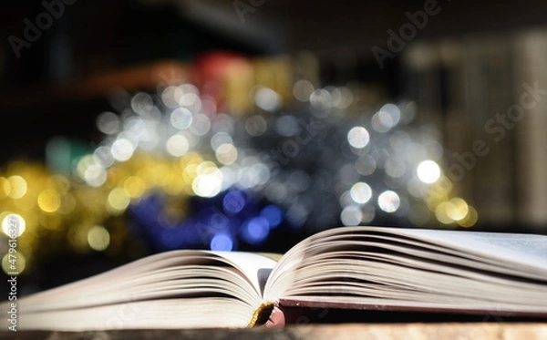 Obraz The open book, illuminated by the sun, lies on the table. Bookshelf background and blurry color spots.