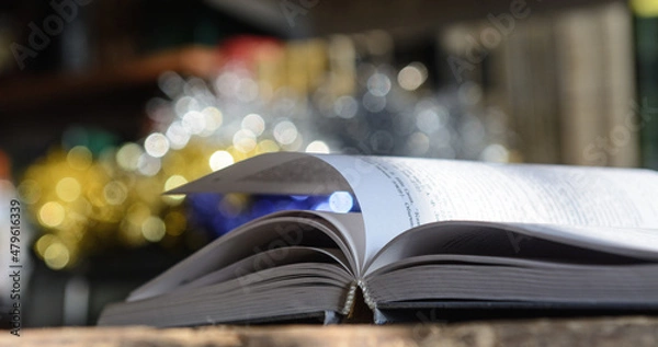 Obraz The open book, illuminated by the sun, lies on the table. Bookshelf background and blurry color spots.