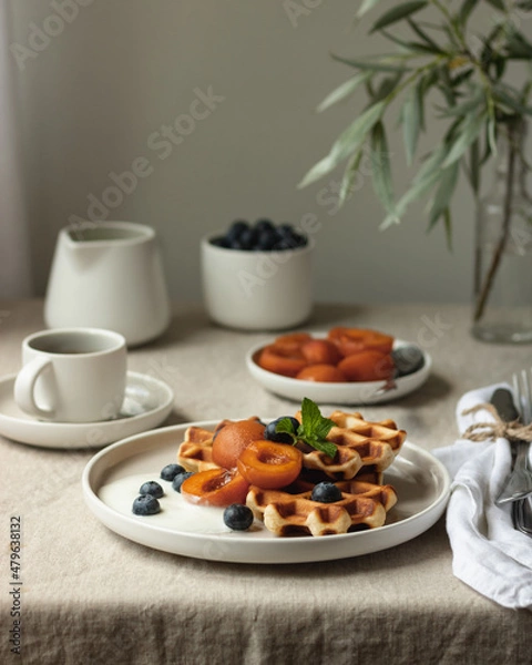 Fototapeta Waffles with addition of fresh blueberries, caramelized apricots and honey on a white plate on table with natural linen tablecloth close up view action shot still life. simply living minimal breakfast