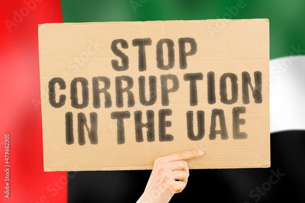 Fototapeta The phrase " Stop Corruption in the UAE " on a banner in men's hand with blurred United Arab Emirates flag on the background. Forbidden. Prevent. Wealth. Offence. Corruptness. Economy. Corruptible