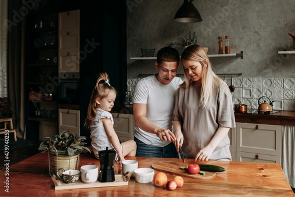 Fototapeta A happy family with one child having breakfast laughing sitting at the dining table in a loft-style kitchen in a cozy house. Pregnant mom dad and little daughter cook together. Selective focus