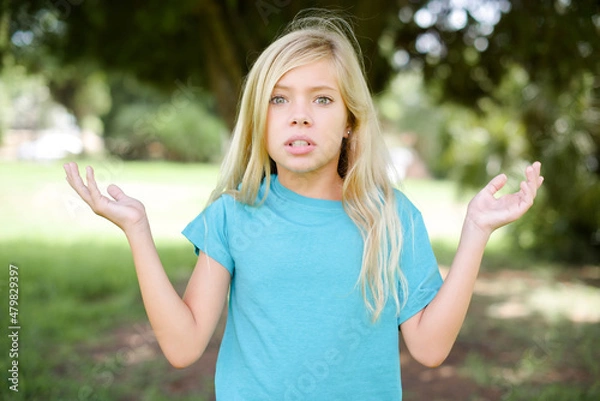 Fototapeta Indignant Caucasian little kid girl wearing blue T-shirt standing outdoors gestures in bewilderment, frowns face with dissatisfaction.