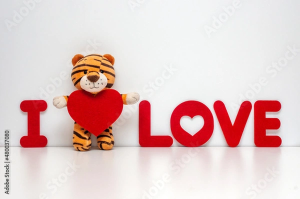 Fototapeta Plush tiger hugging a red heart. A gift for Valentine's Day in the Year of the Tiger. Inscription in red letters I love.