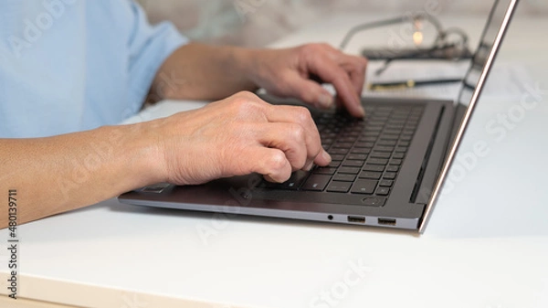Fototapeta Mature middle aged woman using laptop works on the computer while sitting at the table. Senior job seeker looking for a job online. Hands close-up, typing.