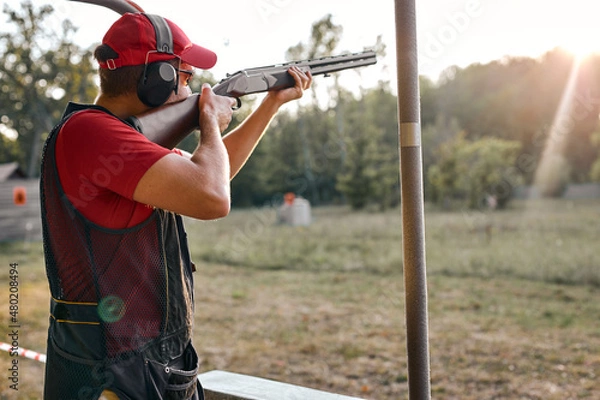 Fototapeta After getting shooting instructions, young caucasian man concentrated at aiming rifle at side, fires at target in outdoor range, wearing goggles, cap and headset equipment outfit. Side view