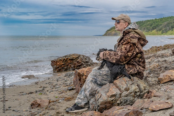 Fototapeta A pensive elderly man by the sea sits on a stone with a dog, thinking about the meaning of life.