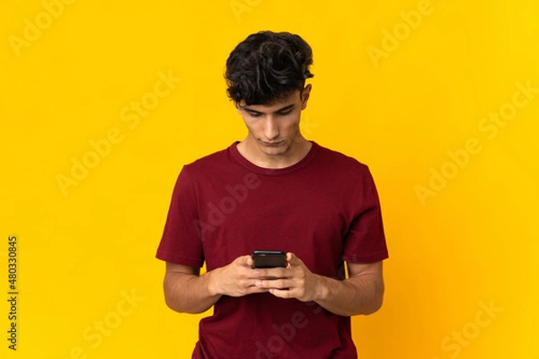 Fototapeta Young Argentinian man isolated on yellow background using mobile phone