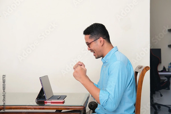 Fototapeta Adult Asian man clenched fist showing success gesture when looking to his mobile tablet screen