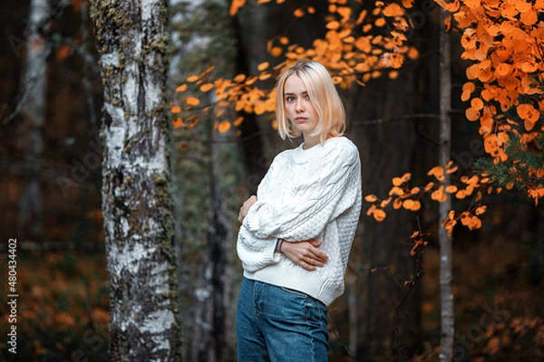 Fototapeta a tall young blonde in an autumn forest dressed in a white sweater and jeans