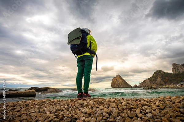 Obraz a girl with a backpack stands on the seashore and looks at the mountains in the distance against the background of a cloudy sky. sunlight breaks through the clouds