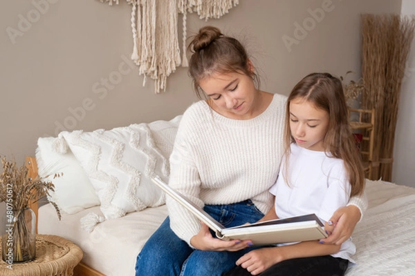 Obraz mother and daughter are sitting at home on the bed flipping through and looking at a book with photos. important moments of life in a photo album. Cute sisters read an interesting fairy tale together
