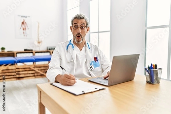 Fototapeta Middle age hispanic therapist man working at pain recovery clinic with laptop scared and amazed with open mouth for surprise, disbelief face