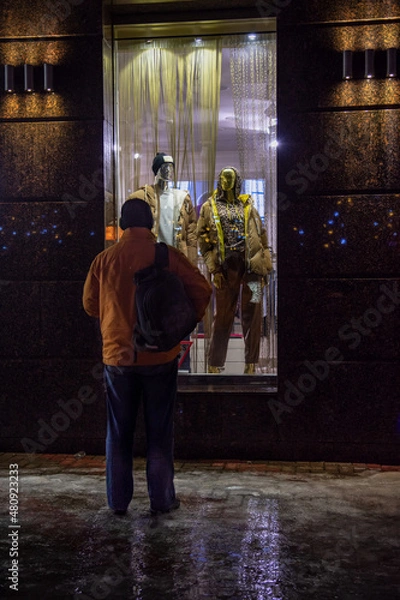 Fototapeta The figure of a man in an old orange jacket and with a backpack in front of a brightly lit window of an expensive fashion store on a dark background