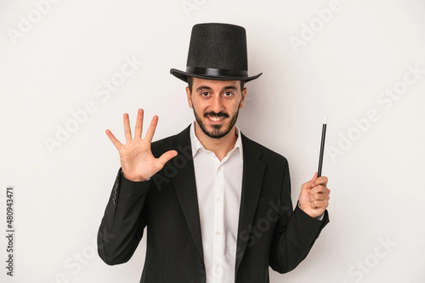 Fototapeta Young magician man holding wand isolated on white background smiling cheerful showing number five with fingers.